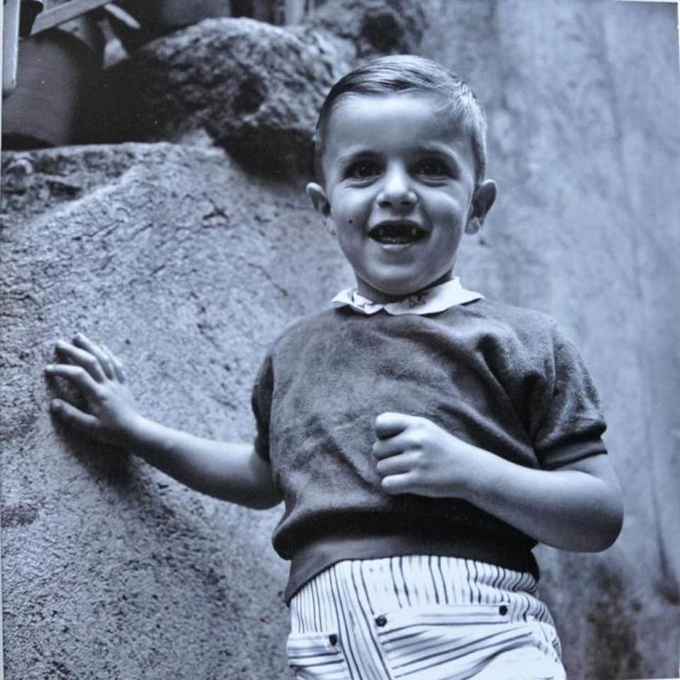 Black and white photograph by Vivian Maier of young Jean-Louis, approximately 4-5 years old, wearing dark sweater with white collar and striped shorts, hands raised mid-gesture with radiant smile, standing against weathered stone wall in Saint-Bonnet-En-Champsaur, 1959